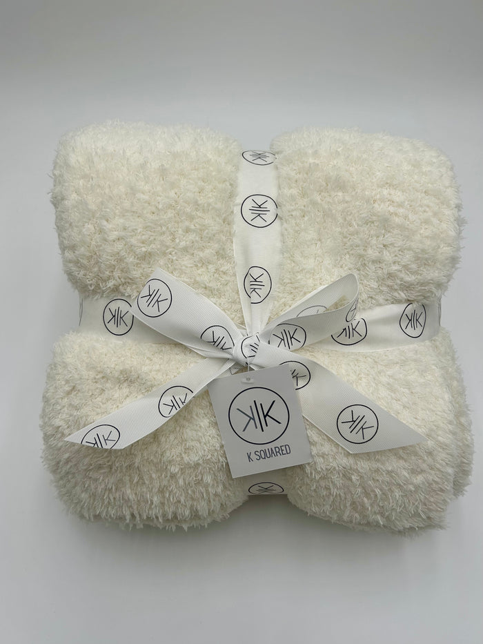 A folded cream-colored fluffy throw blanket tied with a white ribbon printed with the K Squared logo, on a light gray background.