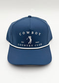 A blue baseball cap with white embroidered text reading 'COWBOY COUNTRY CLUB' and a small golfer graphic on the front, plus a white piping detail around the brim, shown on a light gray background.