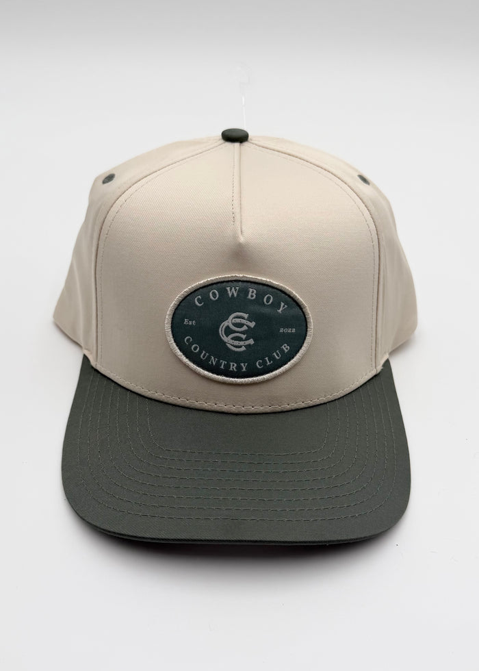 A structured baseball cap with a light beige crown and a dark gray flat brim, featuring an oval patch on the front that reads 'COWBOY COUNTRY CLUB'.
