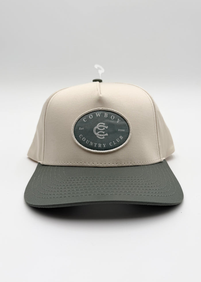 A structured baseball cap with a light beige crown and a dark gray brim, featuring a circular embroidered patch on the front that reads 'COWBOY COUNTRY CLUB'.