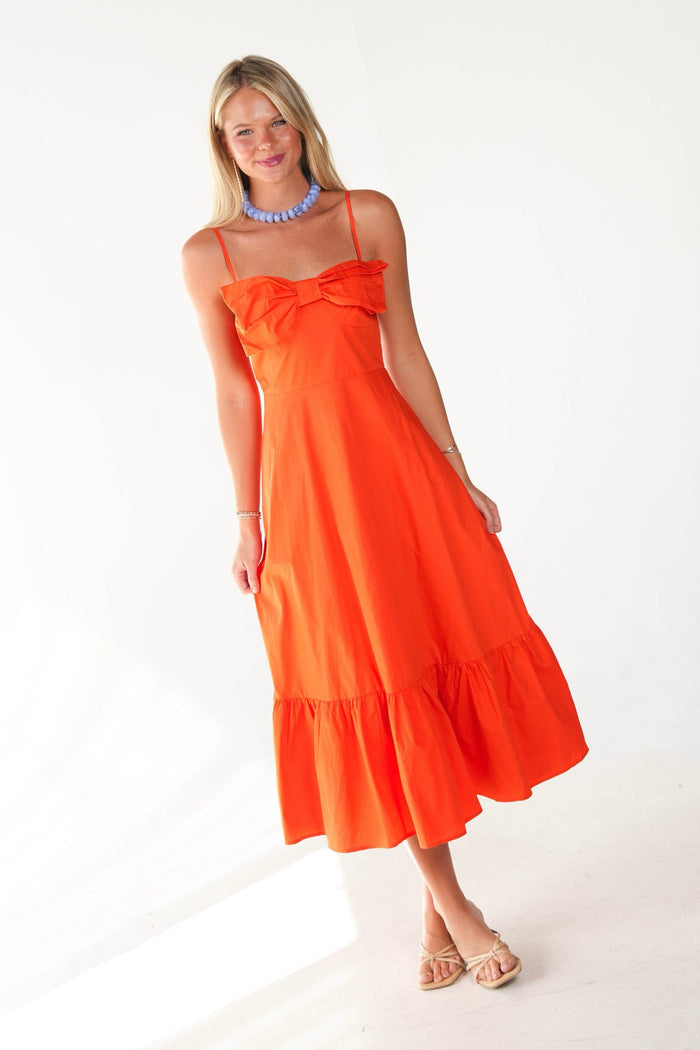 A woman modeling a bright orange midi dress with spaghetti straps, a ruffled sweetheart neckline, and a tiered hem, styled with a blue beaded necklace and beige sandals against a white studio background.