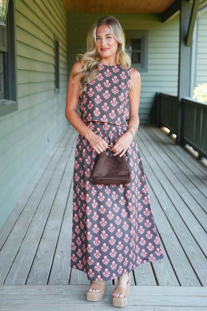 A woman stands on a wooden porch wearing a sleeveless two-piece dress with a pink floral block print on a brown background. She is holding a small brown handbag and wearing beige platform sandals.