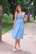 A woman stands on a brick pathway outdoors wearing a blue and white vertically striped halter crop top and matching midi skirt. She is holding a pink and blue pom-pom, with trees and grass in the background.