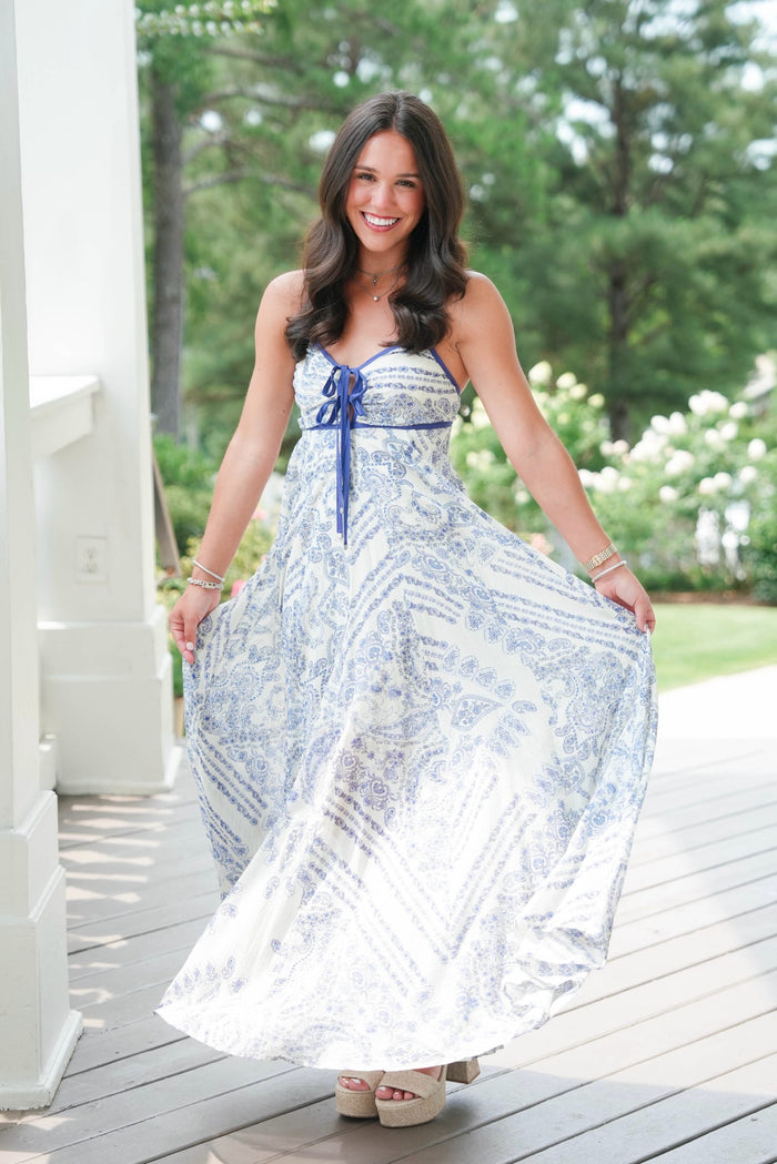 Woman in a blue and white patterned dress standing on a wooden deck with greenery in the background