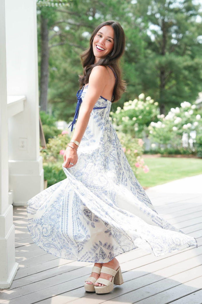 Woman in a white and blue patterned dress standing on a wooden deck with greenery in the background