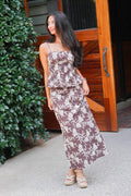 A woman modeling a two-piece matching set featuring a peplum top and a long maxi skirt in a brown and beige floral print, standing outdoors in front of a wooden door and green foliage.