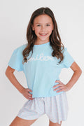 Young girl wearing a light blue t-shirt with 'smile' and white shorts on a plain background