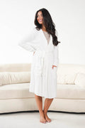 A woman wearing a white long-sleeve wrap robe with a tie waist and front pockets, standing barefoot in front of a cream-colored sofa against a white wall.