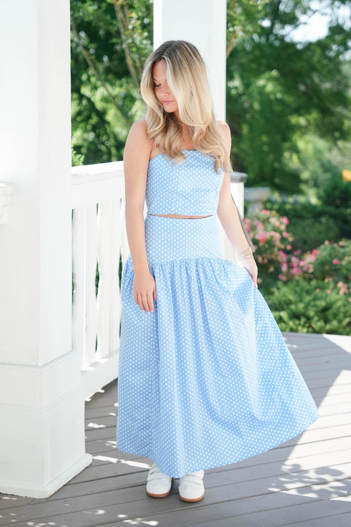 Woman wearing a light blue polka dot dress standing on a wooden deck with greenery in the background