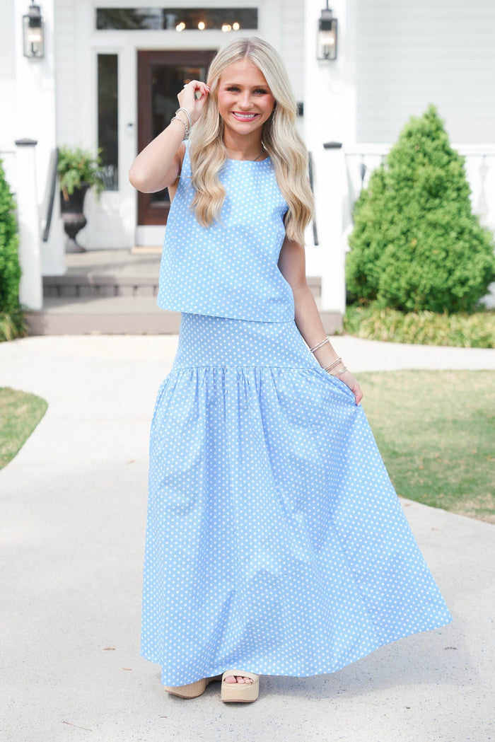 Woman wearing a light blue sleeveless top and long skirt standing outdoors.