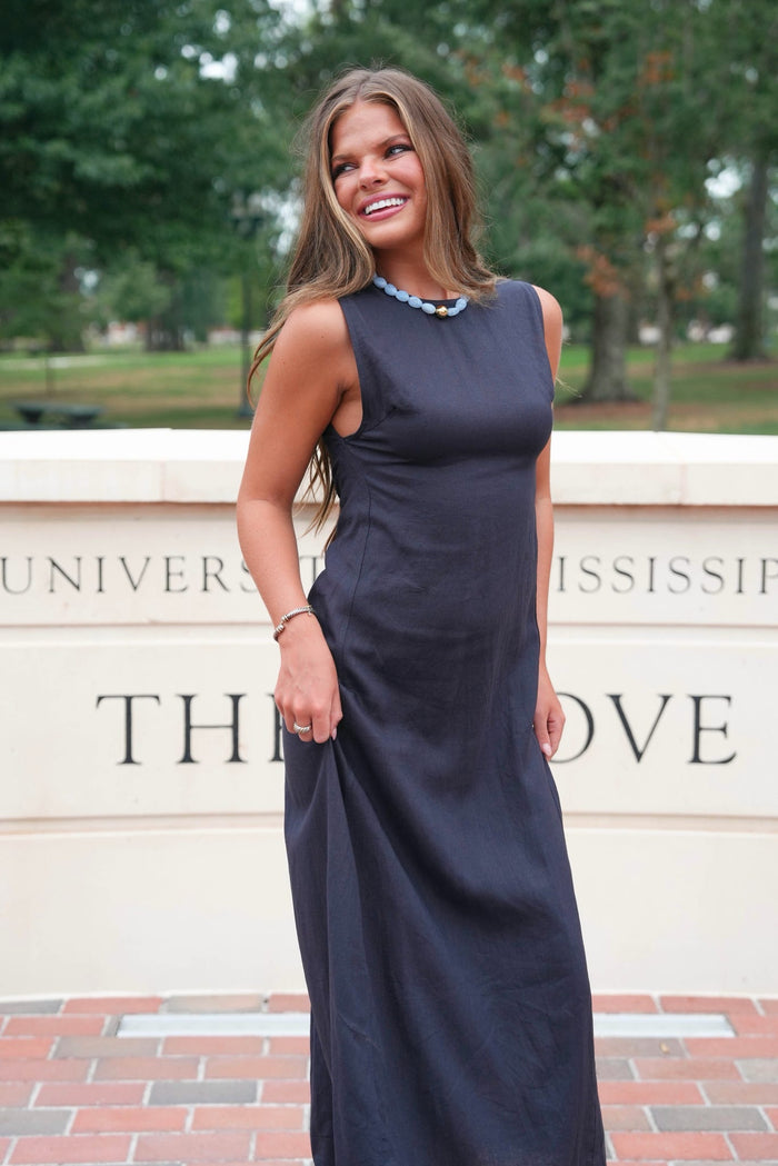 A woman wearing a sleeveless navy blue maxi dress stands outdoors in front of a stone wall with brick flooring. The dress has a fitted bodice and a straight, floor-length skirt.