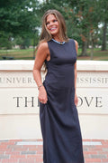 A woman wearing a sleeveless navy blue maxi dress stands outdoors in front of a stone wall with brick flooring. The dress has a fitted bodice and a straight, floor-length skirt.