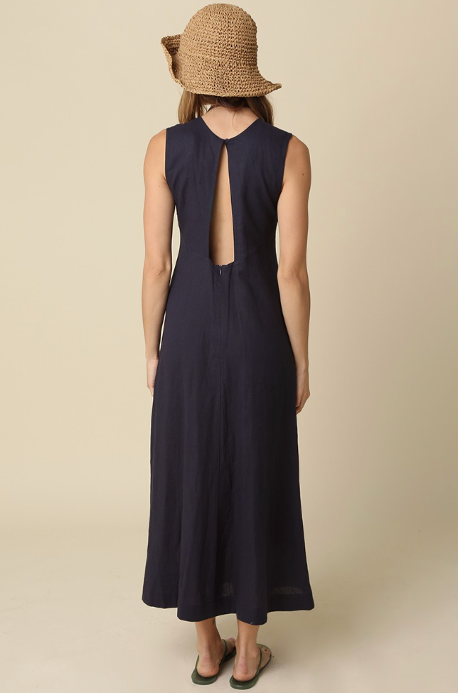 A woman shown from the back wearing a sleeveless navy maxi dress with a large open back and a woven straw hat, standing against a plain beige studio background.