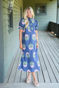A woman stands on a wooden porch wearing a blue midi dress with a pink and green floral pattern, paired with beige platform sandals.