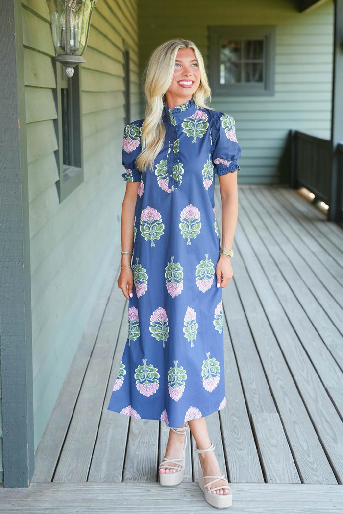 A woman stands on a wooden porch wearing a blue midi dress with a pink and green floral pattern, paired with beige platform sandals.