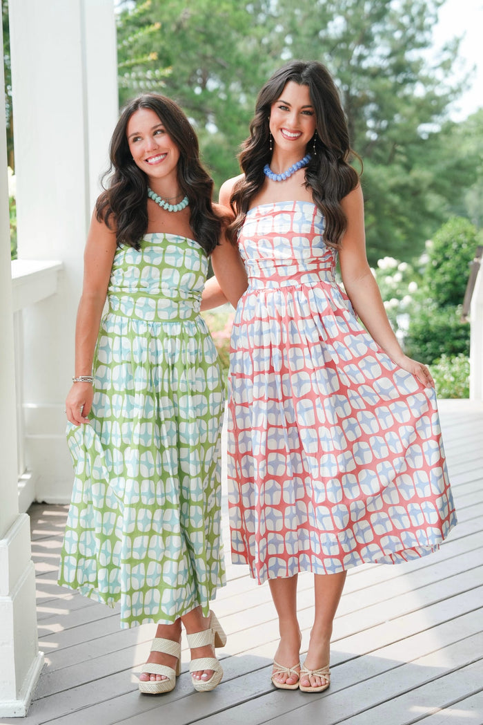 Two women wearing patterned dresses standing on a wooden deck with greenery in the background.