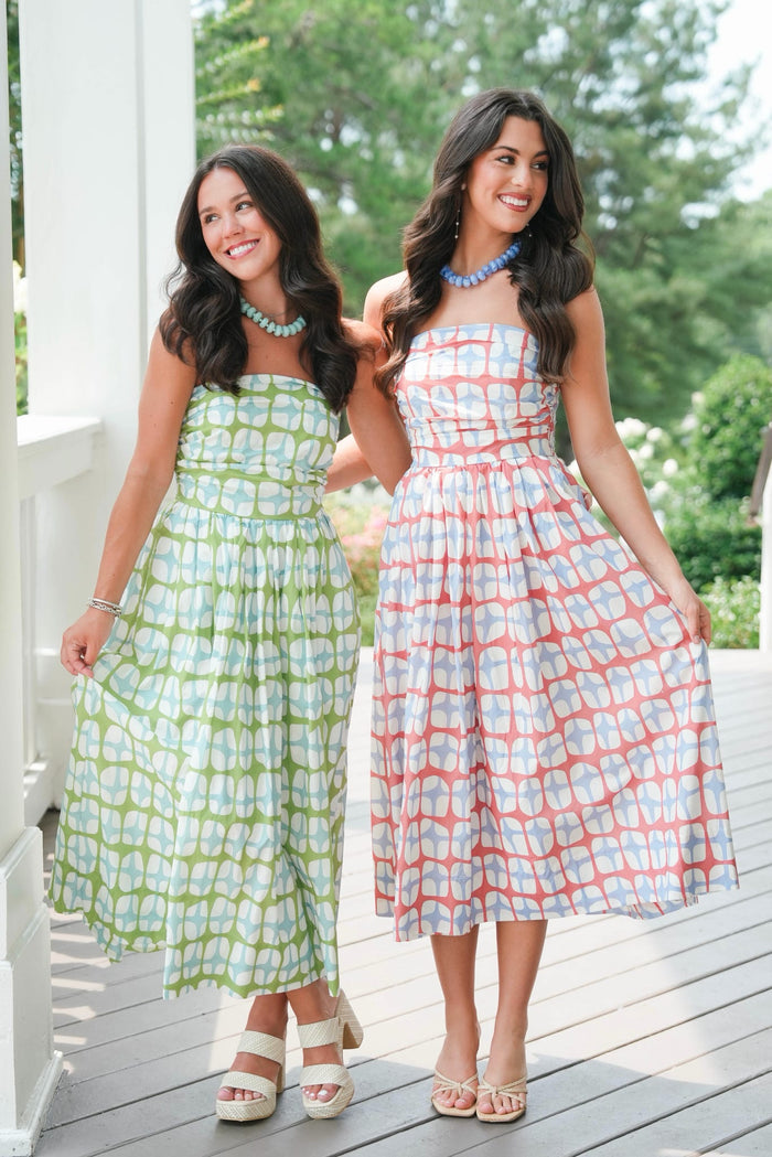 Two women wearing patterned dresses standing on a wooden deck with greenery in the background.