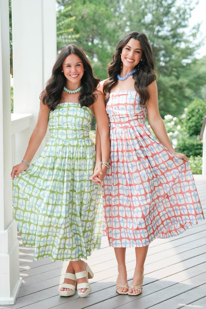 Two women in patterned dresses standing on a wooden deck with greenery in the background