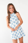 A young girl with long brown hair smiling and wearing a sleeveless white dress with a blue floral pattern, photographed against a clean white studio background.