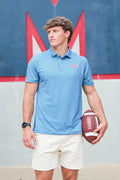 A man wearing a light blue polo shirt with a small red and white logo on the left chest, paired with white shorts, holding a brown football against a blue and red wall background.