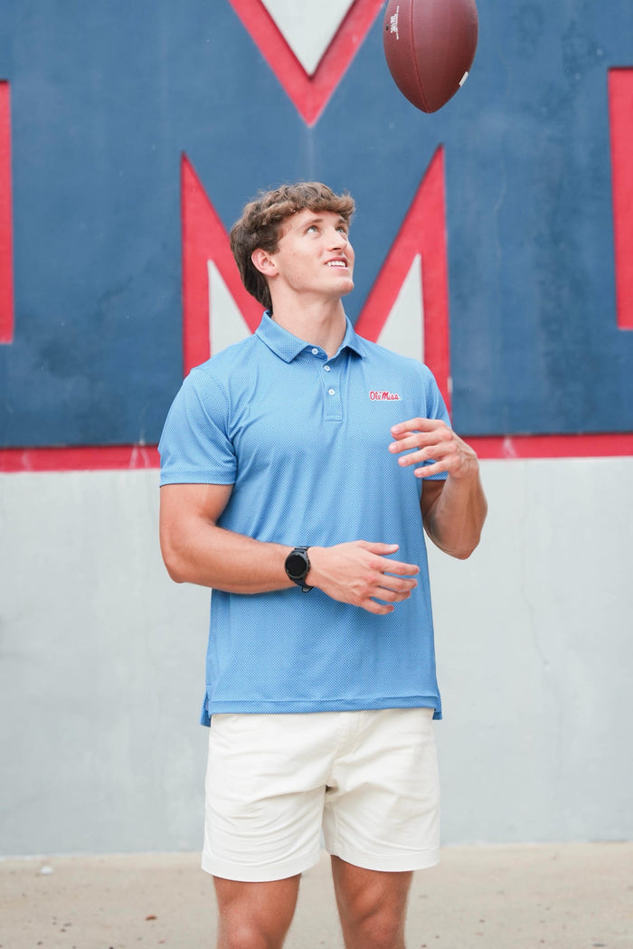 A man wearing a light blue polo shirt and white shorts looks up at a football hanging in the air, set against a blue and red painted wall.
