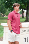 A man wearing a short-sleeve polo shirt in a dusty rose pink color with a subtle geometric pattern, paired with off-white shorts, standing outdoors in front of a stone wall with green trees in the background.