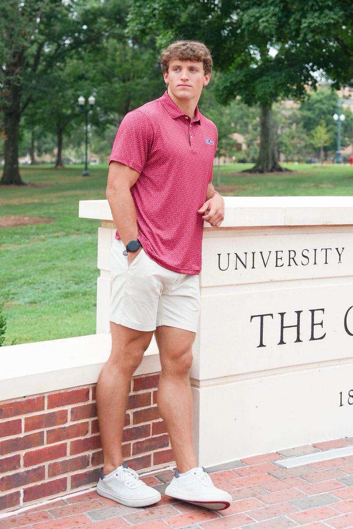A young man stands outdoors leaning against a white stone wall with brick steps below, wearing a short-sleeve textured polo shirt in heathered red, white shorts, and white sneakers. Trees and grass are visible in the background, and part of a sign reading 'UNIVERSITY THE C.