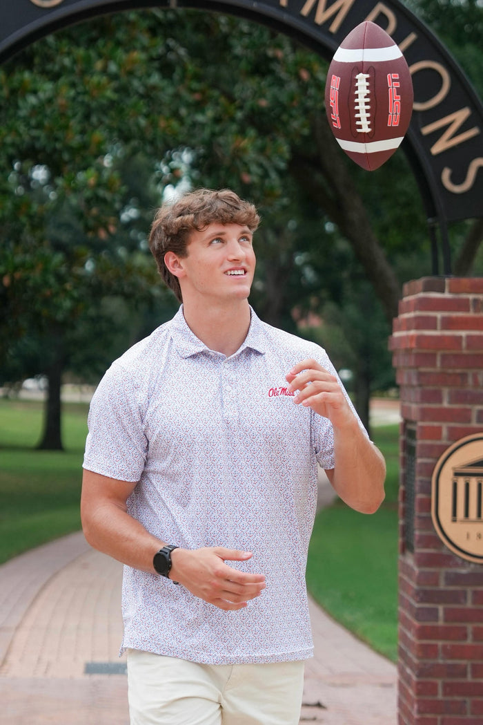A young man in a white patterned polo shirt and khaki pants stands outdoors under a brick archway, looking up at a football floating in the air. The polo shirt has a small red logo on the left chest.