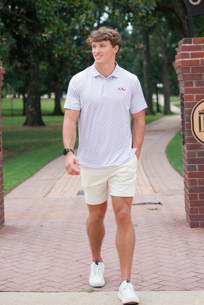 A man walking outdoors on a brick path wearing a white short-sleeve polo shirt with a small red embroidered logo on the chest, cream shorts, and white sneakers.