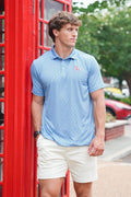 A man wearing a light blue patterned polo shirt and cream shorts stands next to a red telephone booth outdoors.