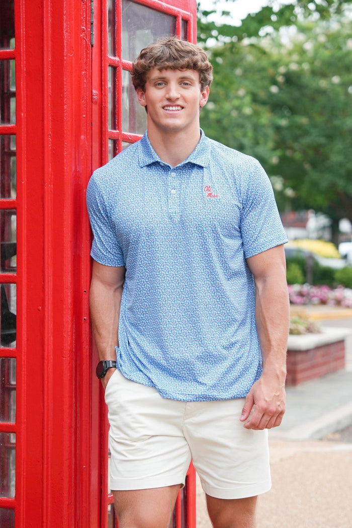 A man wearing a light blue patterned polo shirt and cream shorts stands next to a red telephone booth outdoors.