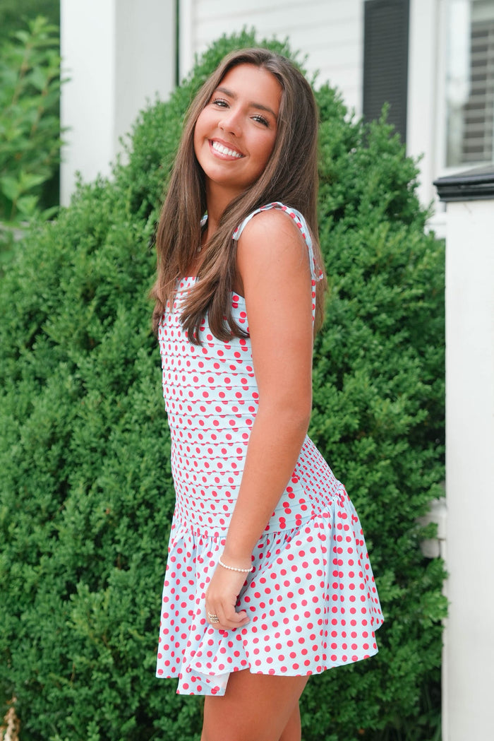 Woman wearing a red and white polka dot dress standing in front of green bushes.