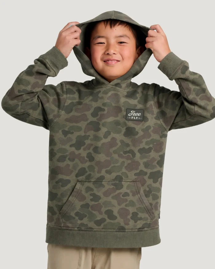 Young boy holding the hood of a green camouflage hoodie over his head in front of a white background.