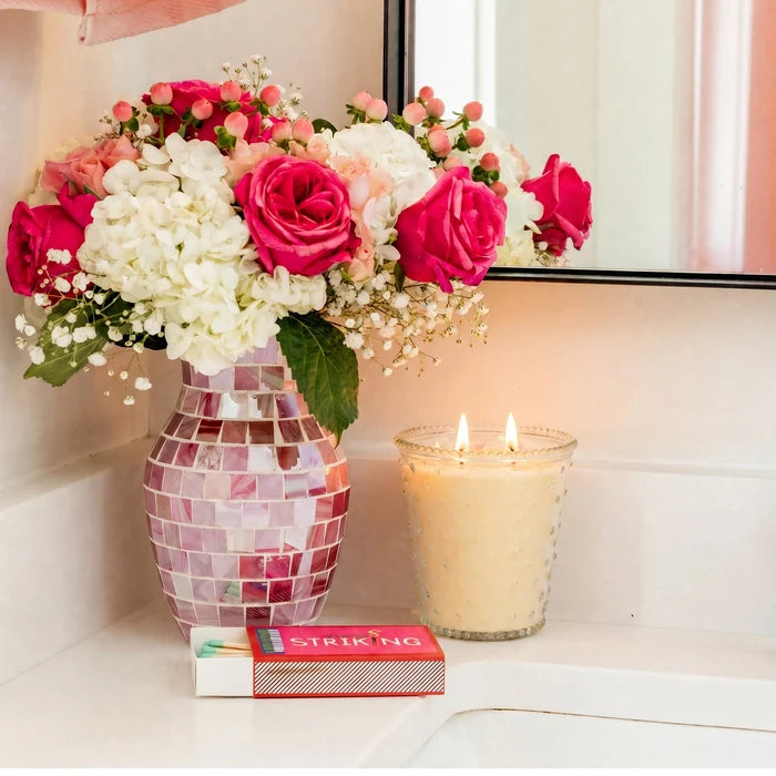 Decorative vase with pink and white flowers on a shelf next to a candle and book.