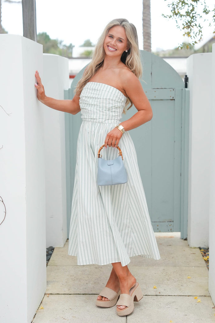 Woman in a strapless white dress holding a blue handbag, standing in front of a white wall and green door.
