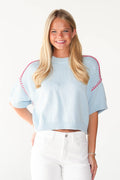 A smiling blonde woman wearing a light blue short-sleeve knit sweater with red contrast stitching along the shoulders, paired with white pants, against a plain white background.