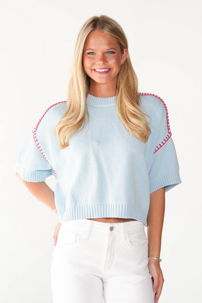 A smiling blonde woman wearing a light blue short-sleeve knit sweater with red contrast stitching along the shoulders, paired with white pants, against a plain white background.