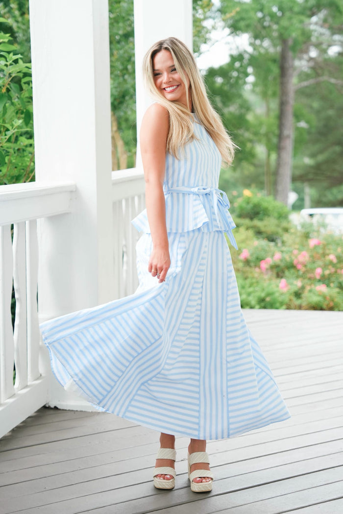 Woman in a light blue striped dress standing on a porch with greenery in the background