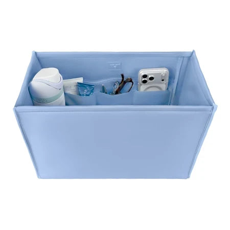 A light blue fabric organizer box with multiple interior pockets, shown holding various small items like a white bottle, a clear pouch, and a white camera. The box is photographed on a clean white background.