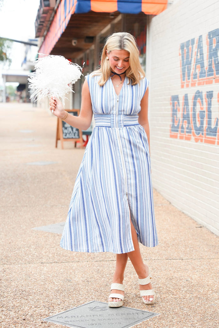 A woman wearing a blue and white vertically striped midi dress with a button-up front and side slit, holding a white feather boa while walking outdoors on a sidewalk next to a brick wall.