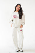 A woman modeling a matching sweatshirt and sweatpants set in a light heather gray color, featuring 'ALABAMA' printed in red on the chest and thigh.