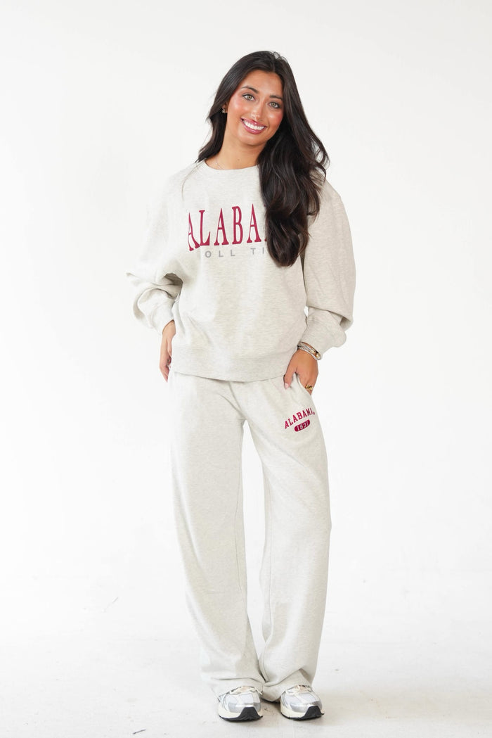A woman modeling a matching sweatshirt and sweatpants set in a light heather gray color, featuring 'ALABAMA' printed in red on the chest and thigh.