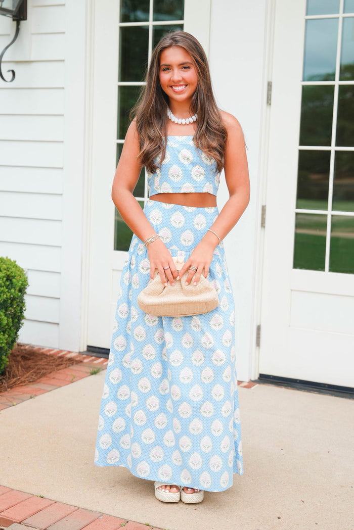 Woman in a blue floral dress holding a beige handbag outdoors.