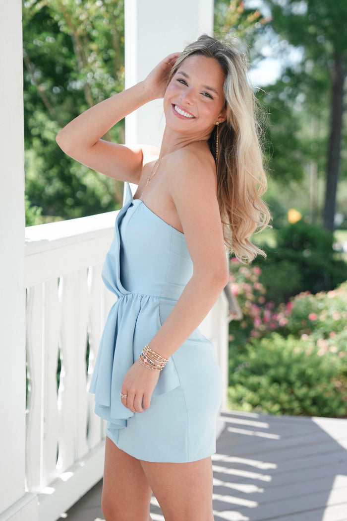 Woman in a light blue dress standing on a porch with greenery in the background