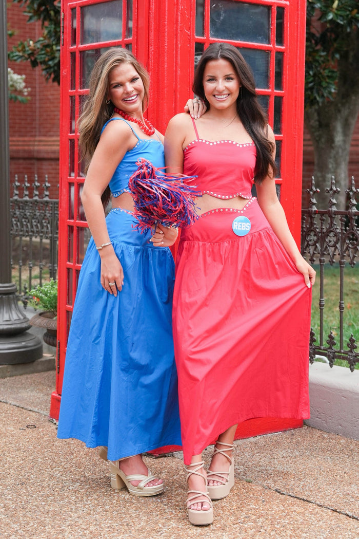 Two women posing in matching two-piece outfits in front of a red telephone booth. One outfit is bright blue and the other is bright pink, both with matching crop tops and long flared bottoms.