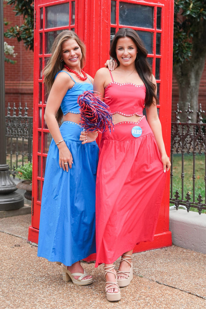 Two women posing in matching two-piece outfits in front of a red telephone booth. One outfit is bright blue and the other is bright pink, both with halter-style crop tops and long flared skirts.