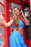 A woman with long blonde hair stands inside a red telephone booth, smiling and holding a black rotary phone. She is wearing a bright blue sleeveless crop top and matching blue skirt with small white decorative accents.