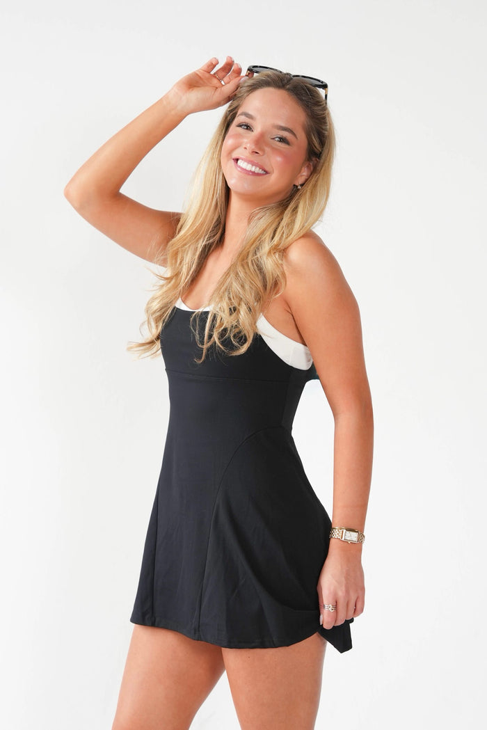 A woman modeling a black sleeveless mini dress with a white band detail at the neckline, posing against a plain white studio background.