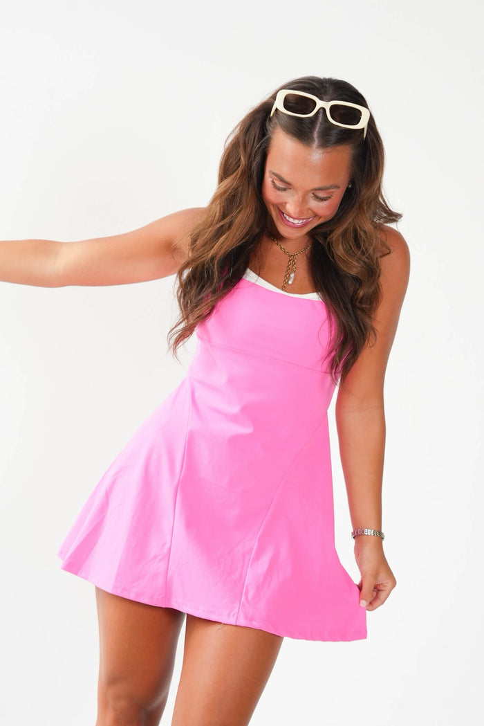 A woman wearing a bright pink sleeveless mini dress with a white inner collar, posing against a solid white background. She has white-framed sunglasses on her head and is smiling.
