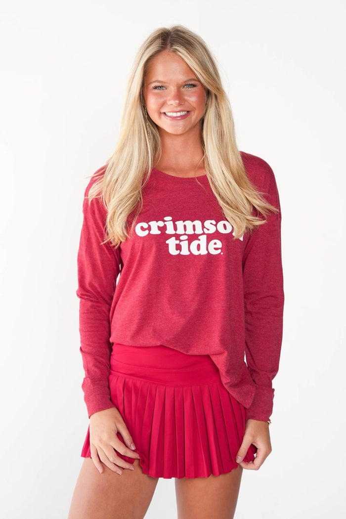 A smiling blonde woman wearing a heathered red long-sleeve crewneck sweatshirt with 'crimson tide' printed in white across the chest, paired with a matching pleated red mini skirt, against a plain white background.